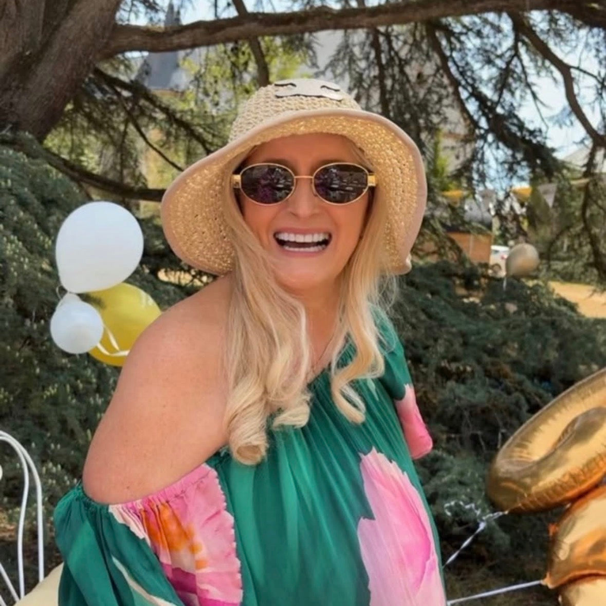 A woman outdoors wearing a straw sun hat, gold-framed sunglasses, and a green off-the-shoulder dress with large pink floral prints. Balloons and trees are visible in the background.